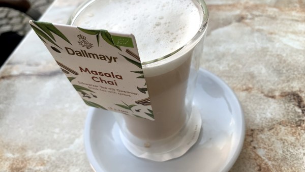 Auf Marmortisch Glas mit Unterteller. In dem Glas ein Milchgetränk mit weißem Schaum. Am Glas ein Schild mit der Aufschrift: Dallmayr - Masala Chai - Schwarzer Tee mit Gewürzen