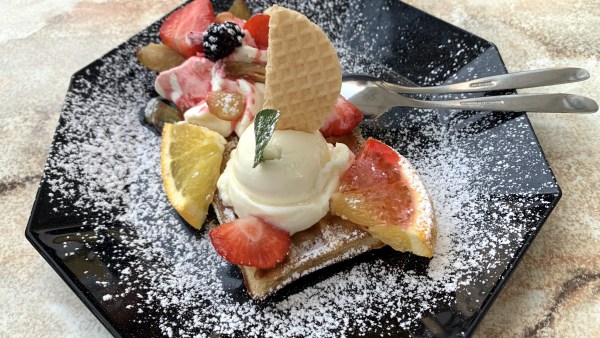 Achteckiger schwarzer Teller auf Marmortisch. Darauf eine Waffel mit einer Kugel Eis und Minzblatt. Dazu Brombeeren, Erdbeeren, etwas Organge. Das Ganze mit Puderzucker bestäubt und mit Löffel und Gabel serviert.