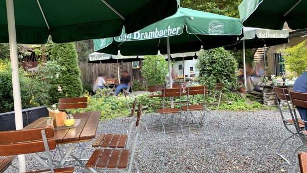 Biergarten: Stühle unter Sonnenschirmen mit Bad Brambacher Aufschrift und Logo auf Kiesboden vor Holzwand und Bäumen. Vereinzelt sitzen Leute. 