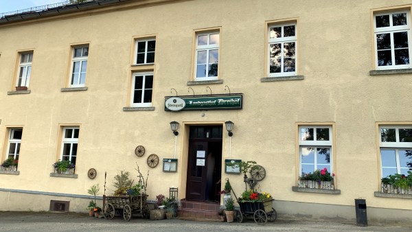 Zweistöckiges Gebäude mit beigem Putz und modernen Fenstern in weißem Rahmen, dekoriert mit Holzwägen und Blumen. Über dem Eingang ein Schild mit der Aufschrift Landgasthof Dreihöf in altdeutscher Schrift und Sternquell-Logo.