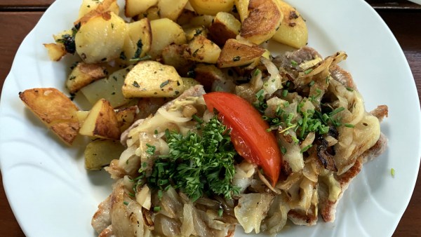 Rostbrätl mit Bratkartoffeln auf weißem Teller, garniert mit Zwiebeln, Petersilie, Schnittlauch und einem Stück Tomate