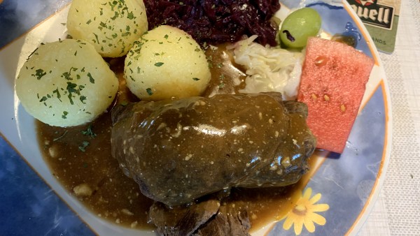 Roulade mit Soße und 3 mit Petersilienkrümel bestreuten Klößen sowie Rotkraut und Weißkraut, einem Stückchen Melone, einer grünen Traube auf einem blauen Teller