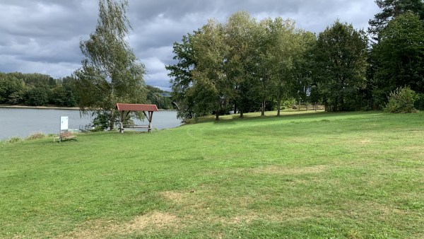 Große gemähte Wiese mit Bäumen am Rand, Sitzecke vor See mitten in Wald bei schönem aber bewölktem Wetter