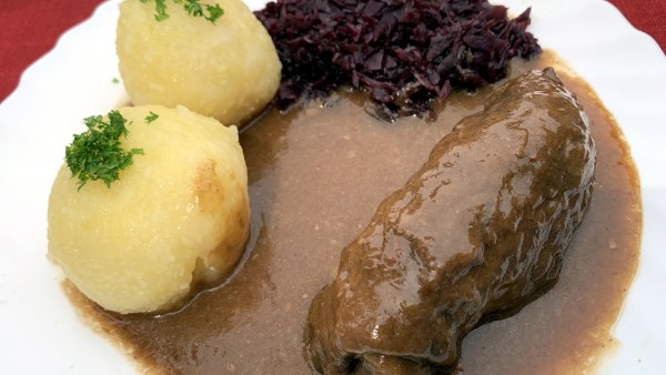 Auf roter Tischedecke weißer Teller mit Roulade, 2 Klößen, Rotkraut und viel Soße. Auf den Klößen ist gestreuselte Petersilie.