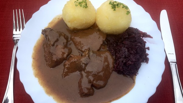 Weißer Teller auf roter Tischdecke zwischen Gabel und Messer. Auf dem Teller 3 Stück Sauerbraten, Klöße mit Petersilie und Rotkraut. Dazu viel Soße.