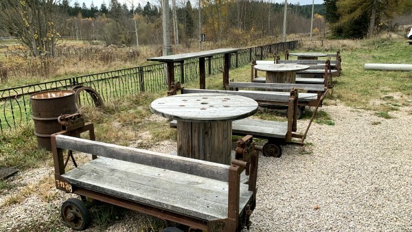 Auf kiesigem Boden vor Wald und Zaun: Holztische und Holzwägen als Sitzmöglichkeiten