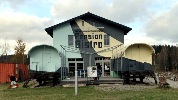 Blau beige und grau gestrichenes Gebäude mit der Beschriftung Weltraumbahnhof Rautenkranz Pension Bistro. Rechts und links je ein halber Eisenbahnwaggon, parallel zum Haus. Alles zwischem orangem Container und Wald. Links steht ein Andreas-Kreuz mit 