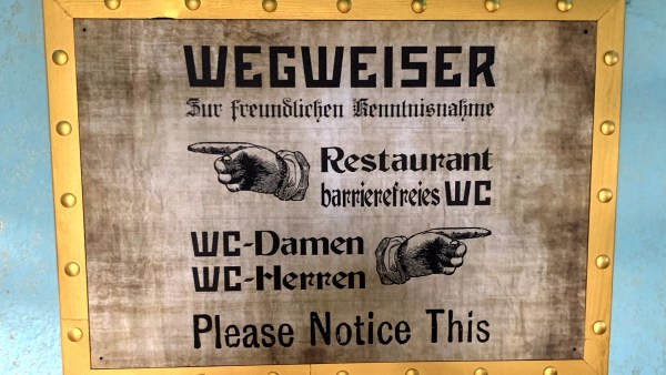 Hinweisschild auf blauer Wand mit vernietetem goldenen Rahmen. Aufschrift: Wegweiser zur freundlichen Kenntnisnahme, Restaurant, Barrierefreies WC, WC Damen, WC Herren, Pleace Notice This