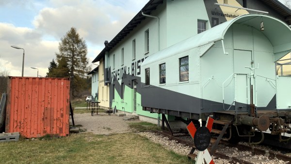 Oranger Container neben blauem Haus. Vor blauem Haus halber blauer Eisenbahnwaggon. Dafor Andreaskreus mit schwarzer Scheibe.